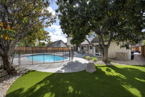 Backyard with grass patch, sitting area under awning, trees, stone pathway, pool with surrounding gate, and enclosing fence